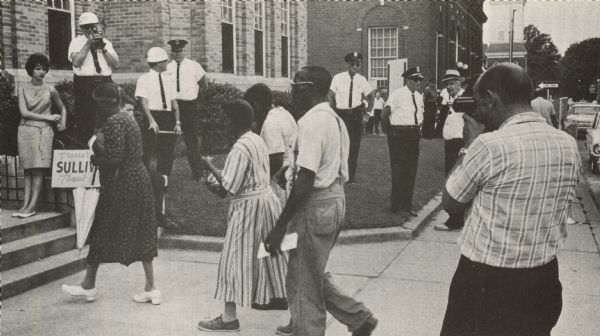 African-Americans approach the courthouse in order to vote. The original caption reads: "Negro citizens attempt to cast ballots in Greenwood, August 1963. Note helmeted policeman and local citizen photographing each of them as they enter the courthouse. The photographs can later be used to intimidate them and perhaps force them from their jobs or homes because they tried to vote."