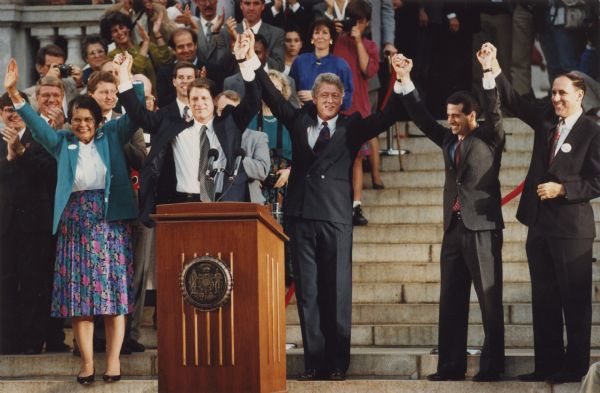 Congressional candidate Ada Deer, on the left, standing behind a podium holding hands with a group of people with their arms raised. The group includes Al Gore, Bill Clinton, U.S. Senate candidate Russ Feingold, and then-Wisconsin Attorney General Jim Doyle. An audience is on the steps behind the group.
