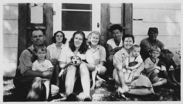 Residents of the Fellowship Farm pose for a casual outdoor group portrait. From left to right are: Walter Uphoff with Gene, Priscilla Stevenson, Dorothy Webster, Betty Wuerth, Bill (Hohri?), Mary Jo Uphoff with Charles, Ernie Hoehner, and Norman Uphoff. 
