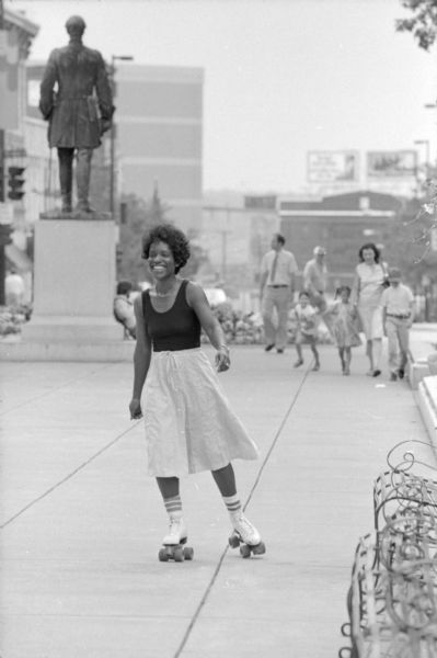 Marlene Cummings, Governor Lee Dreyfus' Advisor on Women and family Initiatives, relaxes by roller skating around the Capitol Square. In the background looking towards King Street is the statue of Hans Christian Heg and pedestrians on the sidewalk.