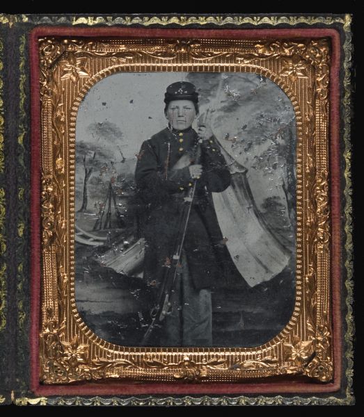 Sixth plate ferrotype/tintype of Ceylon Childs Lincoln as a young soldier in full Civil War uniform. Full-length portrait. He is standing in front of a painted backdrop featuring an army tent. He is holding his rifle with the stock on the ground, and his hands on the barrel near his shoulder. Hand-coloring on cheeks and buttons. 