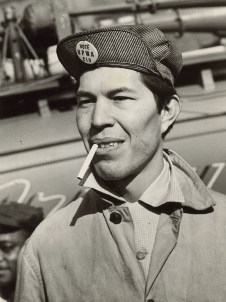 Portrait of a meat packinghouse worker wearing a striped hat. He has an unlit cigarette in his mouth. The button on his hat reads: "Vote UPWA CIO."