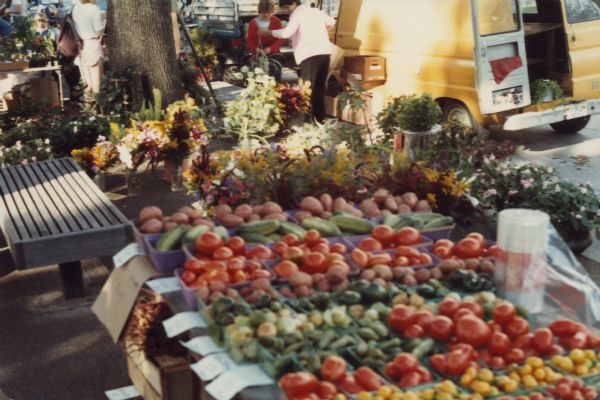 A display of vegetables for sale at the Dane County Farmers' Market. People are standing in the background, and trucks are parked along the curb.