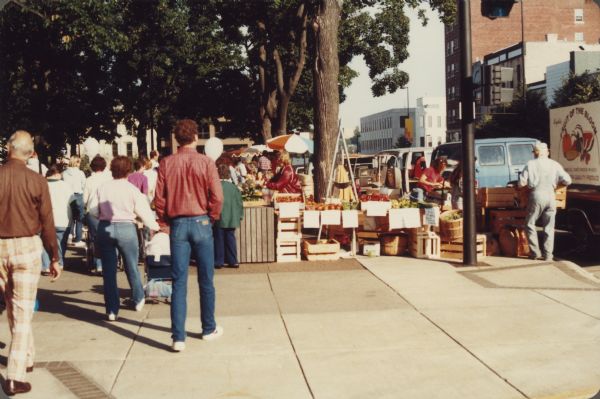 People are strolling along the sidewalk at the Dane County Farmers' Market on the Capitol Square. Men and women are working at their stands, and vehicles are parked along the curb.