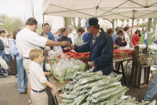 A man is selling produce to customers on the opening day of the Dane County Farmers' Market on the Capitol Square. The Wisconsin State Capitol is in the background on the right.