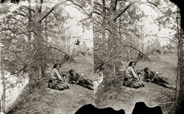 Stereograph of Coo-Nu-Gah (First Boy) and Big Bear, two Ho-Chunk men sitting on the ground. The man on the left is holding a gun.