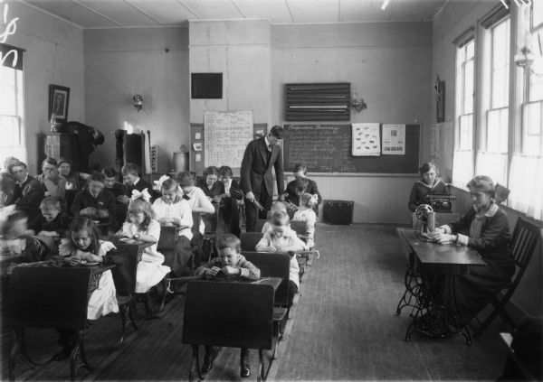 A classroom of students at their desks. There is a woman sitting at a sewing machine and another woman sitting at a typewriter near the windows on the right. At the back of the room the male teacher is assisting a child who has a bundle of grain on their desk. Other students are working with their stalks of grain.