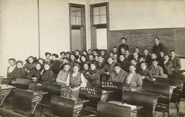 A class portrait of high school students seated at desks. There are two adults, most likely teachers, standing behind them by a chalkboard. Written on the postcard: "High School, Arena, Wis."
