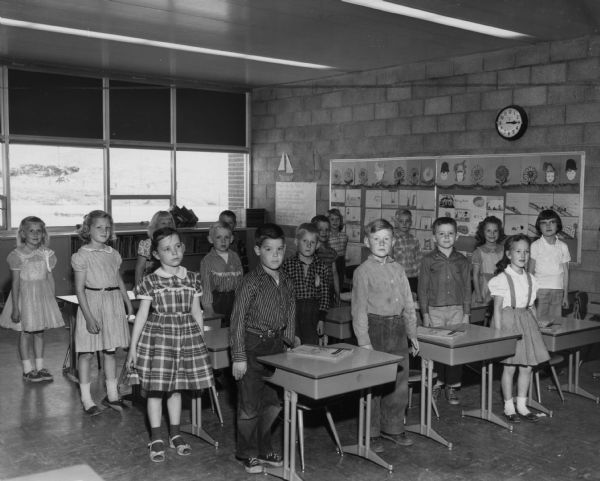 A class of first grade students stand by their desks. 