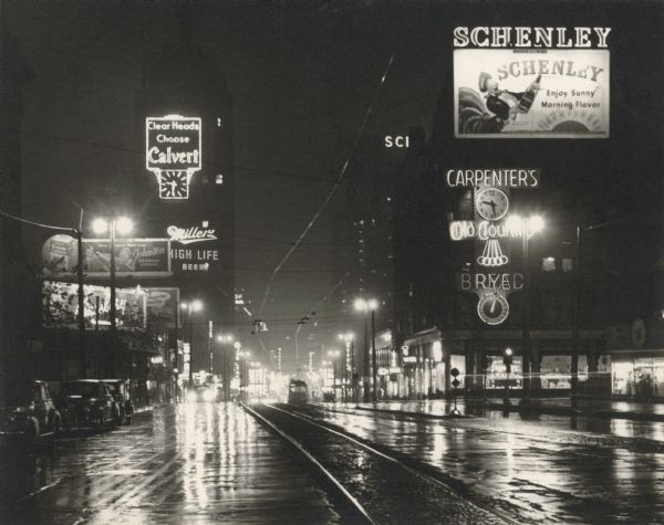 Wisconsin Avenue at Sixth Street, looking east from near the front of the Milwaukee Public Library (left foreground). Taken at night; lighted clocks read 9:30. Lighted  signs advertise Carpenter's Old Country Rye Bread, Schenley (liquor), Calvert (liquor), and Miller High Life (beer). A distant street car is heading away from the camera, and light is being reflected from the tracks and the wet street.