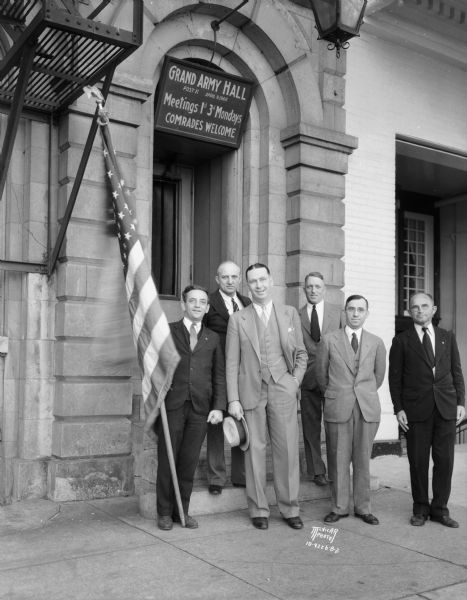Six men with a flag in front of Grand Army Hall, 118 Monona Avenue, (Martin Luther King, Jr. Boulevard). Taken as publicity for the motion picture "Forgotten Man." Includes: John Skagen, Orin Brigham, Horace Goetstein, past commanders, H.E. Hebblethwaite, commander, Edward "Ace" Fischer, incoming commander, and another man. The sign above the entrance reads: "Grand Army Hall, Post 11 April 9, 1866. Meetings 1st 3rd Mondays. Comrades Welcome."