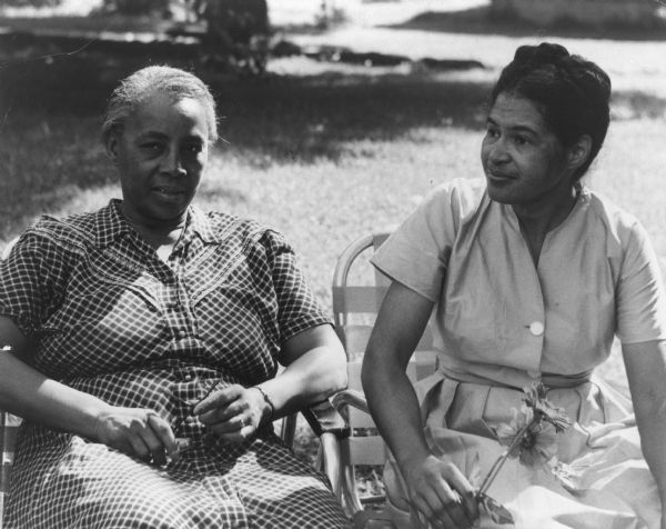 Septima Clark (left) and Rosa Parks are sitting outdoors next to one another. Clark is holding her eyeglasses in her lap, and Parks is holding flowers.