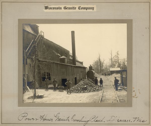 View down railroad tracks toward a group of six men posing outdoors at the power house. There is a pile of large rocks near the group of men at the corner of the power house in the foreground. Larger piles of smaller pieces of rock are behind the building, and also on the right side of the railroad tracks between two small buildings. Another man stands in the background near the curve of the railroad tracks. Caption at bottom reads: "Power House Granite Crushing Plant, Ableman, Wis."