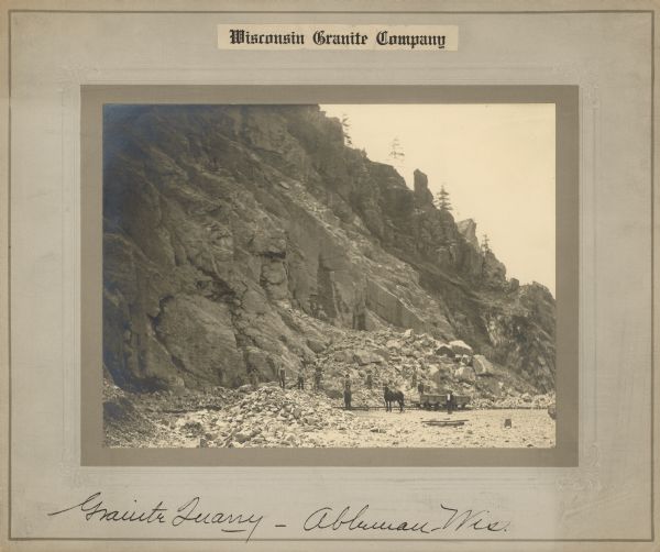 View across the bottom of a quarry toward a group of men posing around a horse who appears to be pulling an open railroad car on railroad tracks. Steep cliffs rise above the group on the left, and piles of large pieces of stone are at the base. Caption at bottom reads: "Granite Quarry, Ableman, Wis."