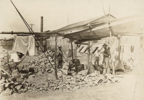 Paving block cutters at work at the the Montello Granite Company. Two men holding large hammers are posing underneath a tarp which is shading large pieces of rock they are working on. On the right behind the tarp are two other men standing behind them. There are piles of smaller pieces of the rock in the yard on the left. In the background is an industrial building and a smokestack.