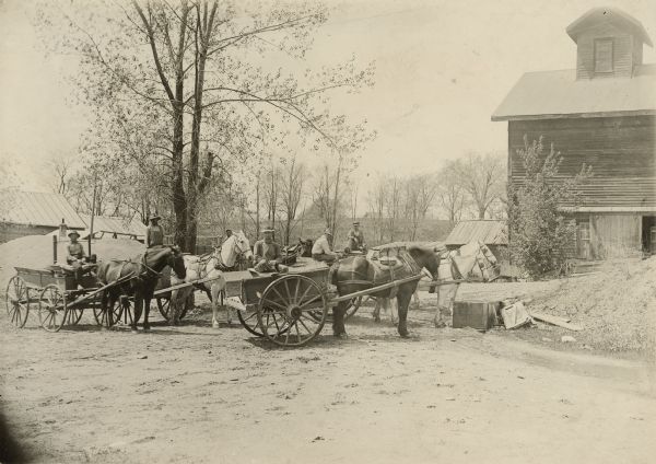 Six men are posing on horse-drawn wagons at quarry houses for the Montello Granite Co. A large building is on the right, and a pile of stone and two small buildings are on the left in the background. In the distance is a hill and trees.