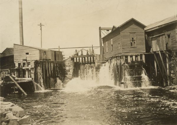 Water power at the Montello Granite Co. View across water toward buildings elevated above the water. Water is pouring through the foundations of some of the buildings, and a gate above a stone foundation is in the center background.