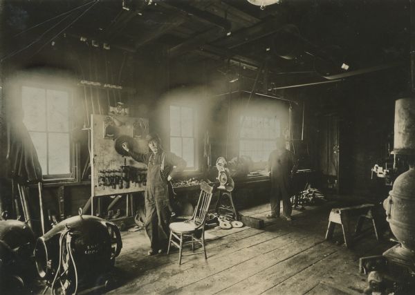 Two men standing inside the machine shop at Montello Granite Co. The man on the left has a pipe in his mouth, and he is standing with his hand on the power supply mounted on the wall. Another man is standing further down the room on the right. Windows are along the wall.