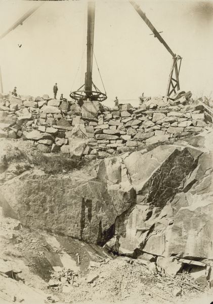 View from below looking up a rockface toward a derrick at the top of a quarry. Men are standing along the top nearby. Caption on back reads: "Quarry view with large derrick on edge of quarry. Montello Granite Co., Montello, Wis."