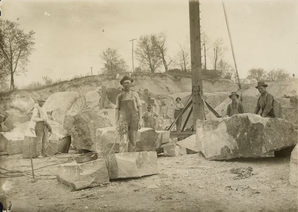 Group of men posing outdoors among large stones. Caption on back reads: "Shaping stone for monuments, Montello Granite Co., Montello, Wis."