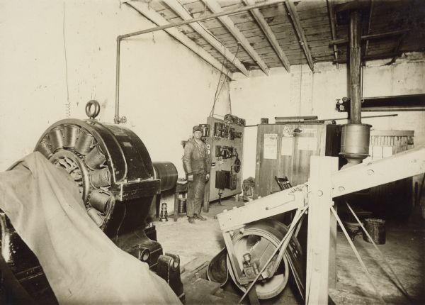 View of the interior of the power house, with dynamo. A man is standing near the wall at left center, near an electrical control panel. Caption on back reads: "Dynamo and Power house, Montello Granite Co., Montello, Wis."