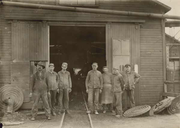 View down railroad tracks toward a group of men posing outside in front of the open doors of a shed. Round polishing stones are laying on the ground on the left and right. The railroad tracks continue into the inside of the shed. Caption on back reads: "Polishing shed and polishers, Montello Granite Co., Montello, Wis."