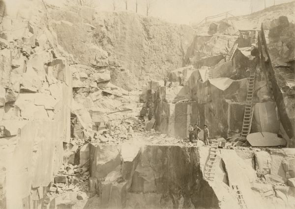 View across the quarry toward three men standing on a ledge below a large rock face. Ladders are set up above and below the ledge. Another group of men are above them on another ledge on the left. Trees are at the top of the quarry in the background. Caption on back reads: "Where the celebrated Montello Granite is quarried. Montello, Wisc."