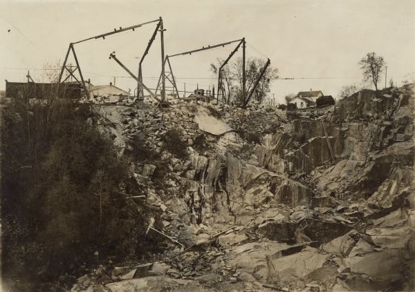 View looking across the quarry, which has trees and plants growing up the sides on the left, and smaller rocks and other debris are toward the bottom. Ladders and wheelbarrows are scattered about the quarry. Derricks and other equipment are set up along the far edge, and behind them is an industrial building in the background on the left. Smaller buildings are in the background on the right. Caption on back reads: "Scene in the quarry. Montello Granite co., Montello, Wis."