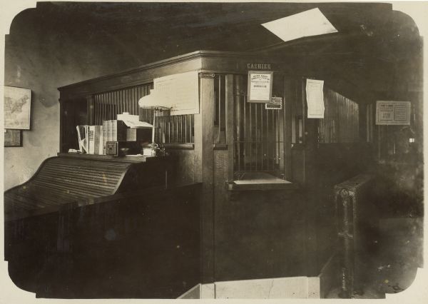 Bank interior. On the left is a roll-top desk. There is a sign for "Cashier" on the wall in the center at the barred window. A framed certificate below it reads, in part: "Insured Against Burglary and Hold-Up by Maryland Casualty Co. Baltimore, MD."