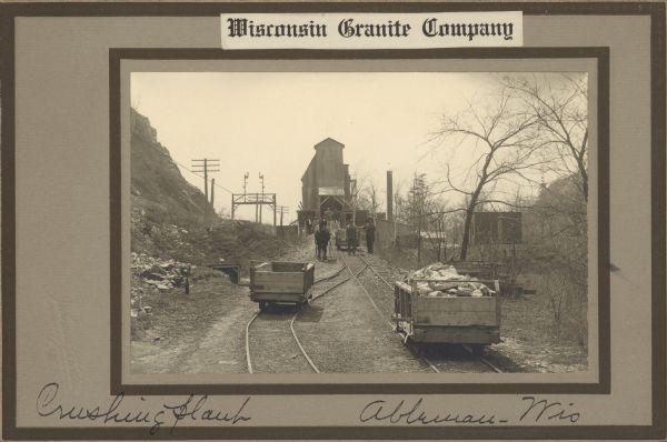 View down two sets of railroad tracks. There is an empty cart on the left set of tracks, and one cart full of large pieces of stone on the right set of tracks. The tracks converge further down, where a horse stands to the side of the tracks pulling another empty cart down from the tall industrial building in the background. Two men are standing nearby, one holding the reins to the horse, and the other holding the side of the cart. Other men are standing behind them on the ramp that leads up to the building. A steep hill is on the left, and trees are along the right. Caption at bottom reads: "Crushing Plant, Ableman, Wis."