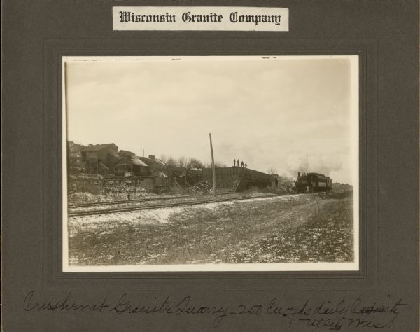 Angled view across two sets of railroad tracks toward industrial buildings along a hill on the left. A locomotive pulling a railroad car is coming down the tracks from the right. Men are standing on top of the wall of a building or large holding bin in the center. Caption at bottom reads: "Crusher at Granite Quarry — 250 Cu yds daily capacity, Utley, Wis."