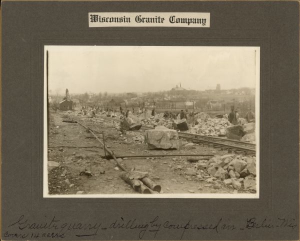 View towards railroad tracks and men working with large pieces of stone. In the distance is the town of Berlin. Caption at bottom reads: "Granite Quarry — drilling by compressed air — Berlin, Wis."