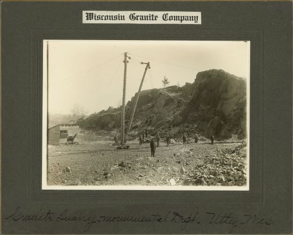View towards men standing around an open area. Two tall poles with pulleys near the top is supported by wires are in the center near a set of railroad tracks. Behind them is a large rock formation at the quarry. A small industrial building is on the left. Caption at bottom reads: "Granite Quarry — Monumental Dept. — Utley, Wis."
