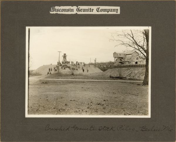 View across open field toward a large group of men posing on a large pile of granite. There is a large industrial building in the background on the right. Caption at bottom reads: "Crushed Granite Stock Piles — Berlin, Wis."