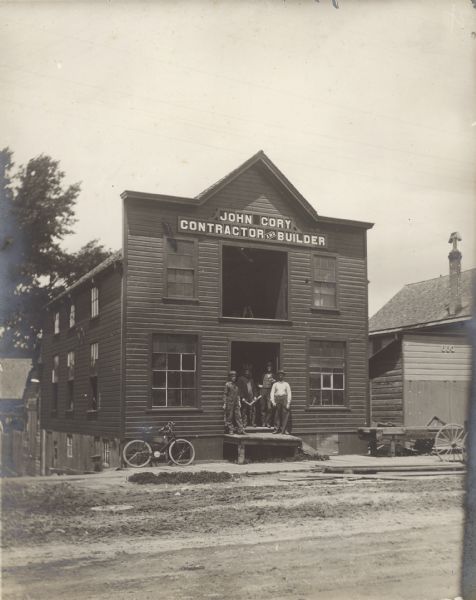 View across unpaved street toward a group of four men standing on a platform in front of the large open door of a building. A larger open door is above them on the second floor. Three of the men are holding tools, and a bicycle is leaning against the left side of the building on the board sidewalk. The sign above the second floor door reads: "John Cory Contractor and Builder."