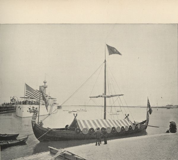 View of a replica Viking Ship that sailed from Norway to the United States for the Columbian Exposition in 1893. Three men are standing on shore in front of the vessel. Another ship is at a dock in the left background.