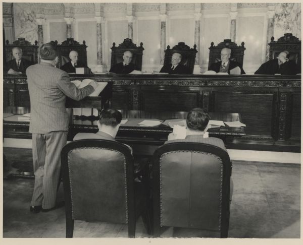 A lawyer presents his case before the Wisconsin Supreme Court. Two other men in the foreground are seated facing the justices.