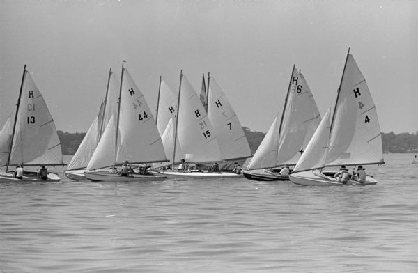 Nine sailboats gather on Lake Mendota searching for wind during the Mendota Yacht Club's racing series. Bill Mattison, Class C; Jim Henkel, Class C; and John Beck, Class X; scored victories in the races. A boat is behind the sailboats, and the sign painted on the front reads: "Courtesy Boat."    