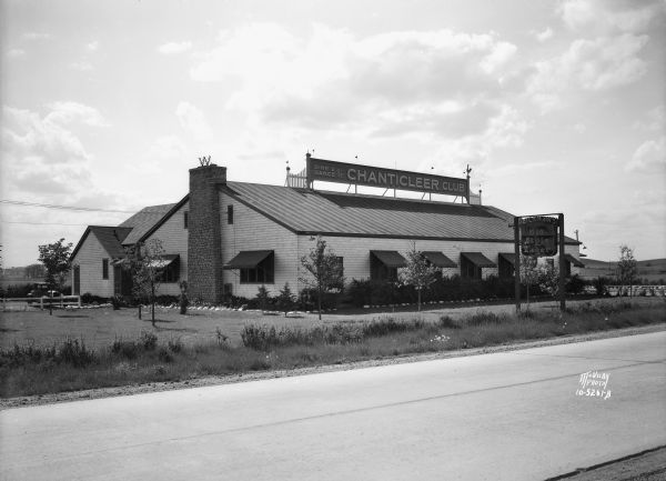 Club Chanticleer, exterior view across road from the south, with a Heileman's Old Style Lager beer sign suspended from posts on the lawn in front. The sign on the top of the building includes the shape of a rooster and small pennants and reads: "Dine & Dance at the Chanticleer Club." On top of the stone chimney on the left end of the building is a small sign with the letter "W." An automobile is parked near a fence on the left near a side entrance.