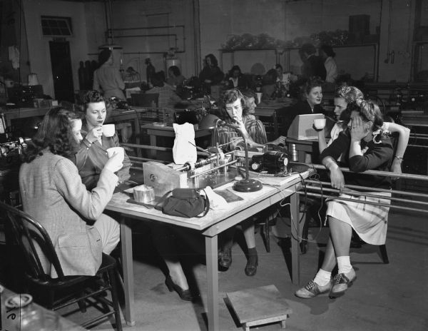 Five women are sitting around a table with machinery and various pieces of equipment. Three of the women are drinking coffee, and one woman is smoking. Women are sitting and standing in the background. Caption reads: "Lake Geneva, Wis. 1943. Women workers at Chaney Instruments. 'Coffee Break.'"