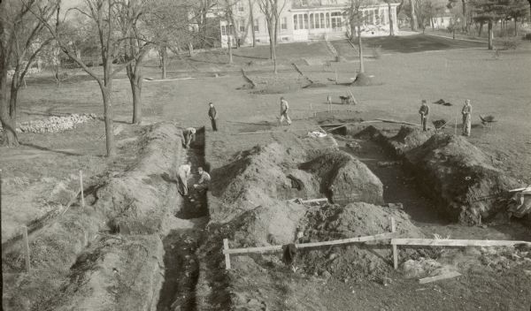 Elevated view of excavations of the first Fort Crawford by Leland Cooper. Seven people are working around the trenches. Villa Louis is in the background.