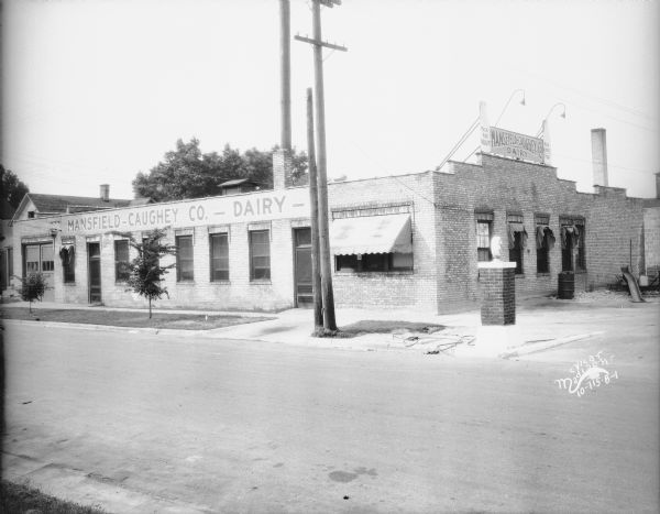 View across street toward the Mansfield Caughey Company Dairy, 701 Williamson Street. The signs on the two milk bottles framing the company sign on the right side of the building read: "Milk For Health" and "Milk A Perfect Food." A sign on a column at the corner reads: "For(?) Gas(?)."