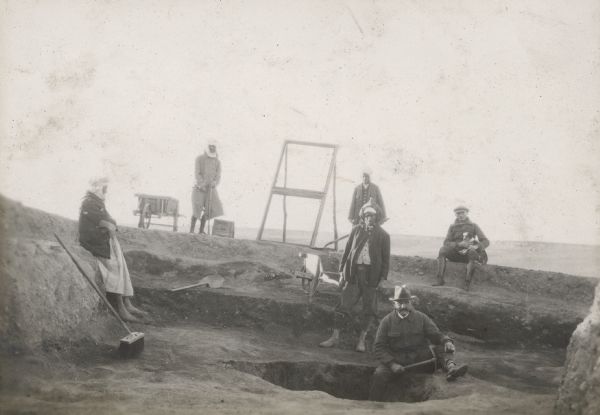 Six people, including Alonzo Pond (standing in the hole on the right side), work at an archaeological dig site in Algeria. The diggings were worked in levels. The man sitting at the top of the trench on the right is holding a dog in his lap.
