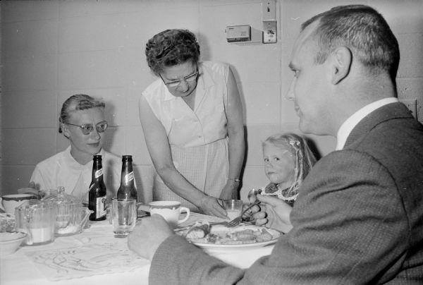 A man, two women and a child are gathered around a table eating a fish fry. One of the women is standing in the center holding a glass of milk.