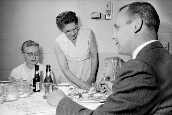 A man, two women and a child are gathered around a table eating a fish fry. One of the women is standing in the center holding a glass of milk.