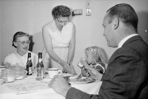 A man, two women and a child are gathered around a table eating a fish fry. One of the women is standing in the center.