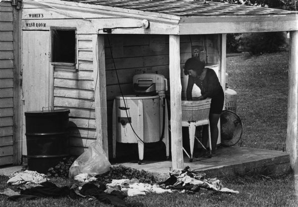 Linda Luna of Auraura, Illinois washes clothes at a basin outside a cabin in a migrant worker camp in Door County. Several articles of clothing are lying in the grass in the foreground, and there is an electric wringer near the basin.