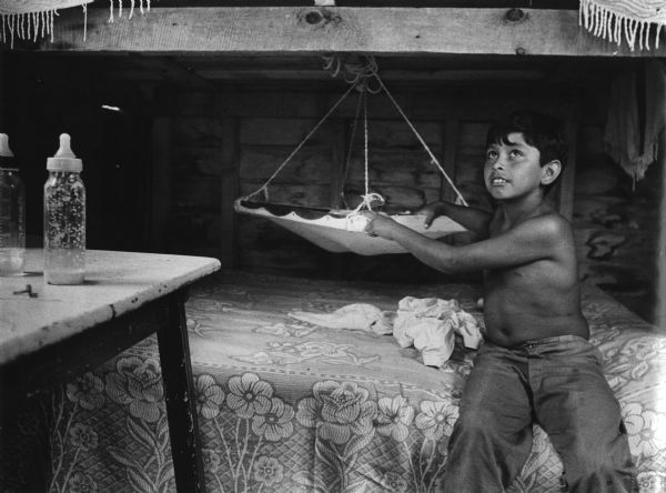 Alberto Herrera watching his baby brother who is in a cradle suspended between bunk beds in a migrant worker cabin near Sister Bay. Two baby bottles sit on a table in the foreground.
