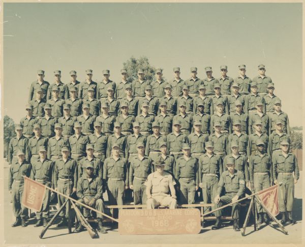 Group portrait of men posing outdoors, including Fernando Rodriguez. The sign in front reads: "Platoon 3065 U.S. Marine Corps, San Diego, 1968, U.S. Marines ••Semper Fidelis••." Flanking the sign are flags that are resting on top of stack arms. Written on back of photograph: "4th row, 6th from left" and "Bootcamp grad 1968 2 drill instr's missing."