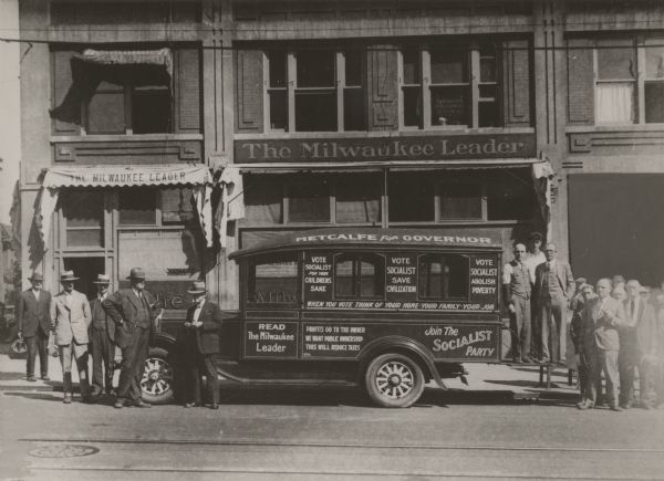 "Metcalf for Governor" is painted on a truck for the campaign. Other phrases: "Vote Socialist For Your Children's Sake," "Vote Socialist Save Civilization," "Vote Socialist Abolish Poverty," "Read The Milwaukee Leader," "Profits Go To The Owner We Want Public Ownership This Will Reduce Taxes," and "Join the Socialist party." The truck is parked in front of "The Milwaukee Leader." Seventeen men surround the truck.